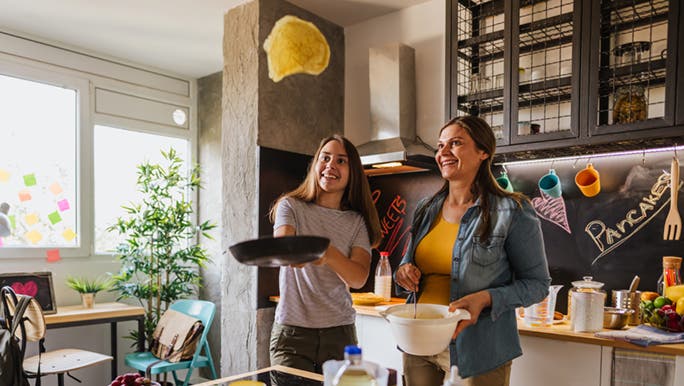 A teenager is flipping a pancake in the air, her mum watches on with an entertained look on her face.