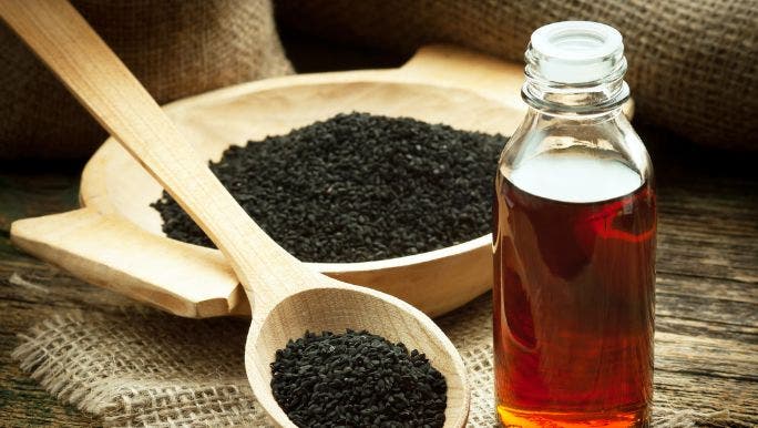 A wooden bowl and spoon filled with black seeds on a table beside a glass jar filled with black seed oil