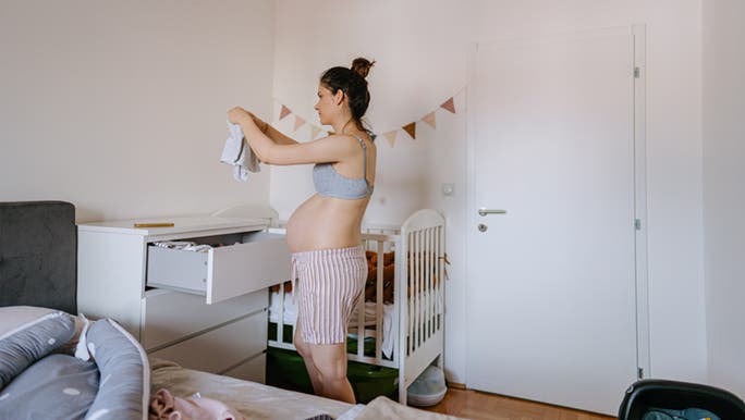A pregnant woman peacefully folds tiny clothes into a set of drawers in a minimalistic nursery.