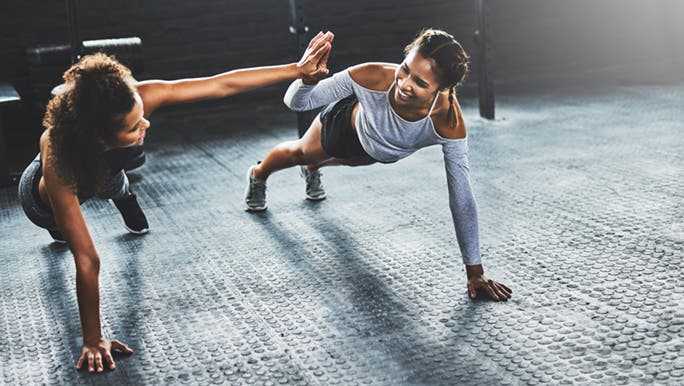 Two friends are high fiving while doing planks together at the gym which is one fitness challenge idea for friends. 