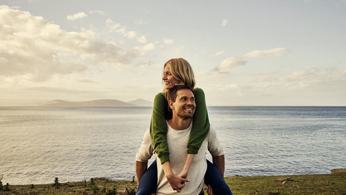 A man is piggybacking a woman at the waterfront, they look happy in their lives. 