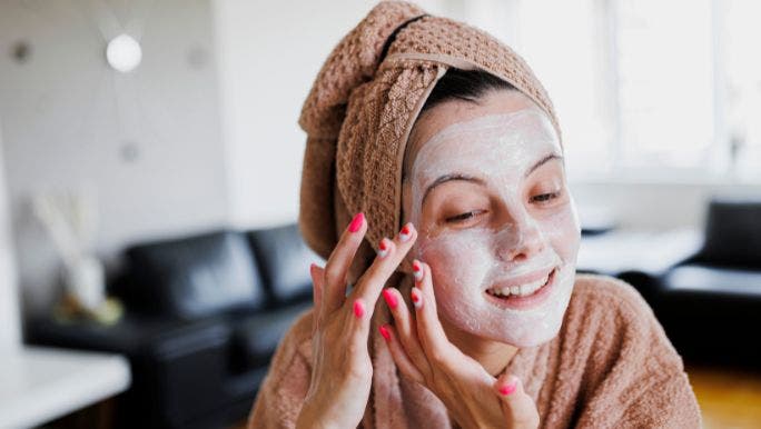 A young woman with bright pink nail polish and her hair wrapped in a towel is applying a skincare mask made of bentonite detox clay