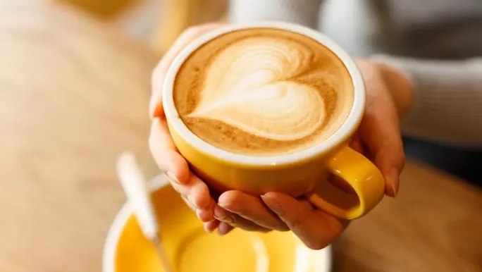 Freshly brewed flat white in a yellow coffee mug being cupped in a woman's hands