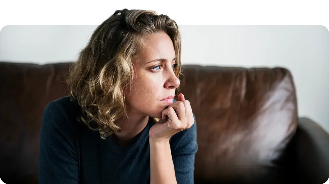A woman is sitting down on a dark brown leather sofa. She is leaned forward and has her chin resting in the palm of her hand. Her eyes are looking into the distance in a concerned manner.