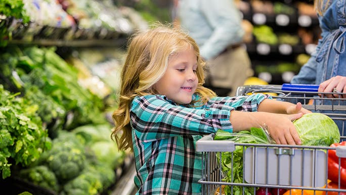 Little girl helping her female caregiver shop for produce in the grocery store, she is putting an iceberg lettuce in the trolley.