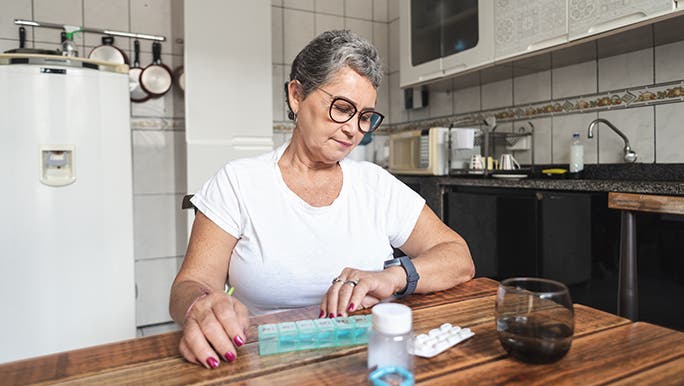 Woman with short salt and pepper hair, glasses and hot pink nails sitting at a wooden kitchen table. There is a pill box and drink on the table, she is looking at her watch. 