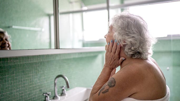 A senior woman is looking at herself in the bathroom mirror as she conducts a good skin care routine