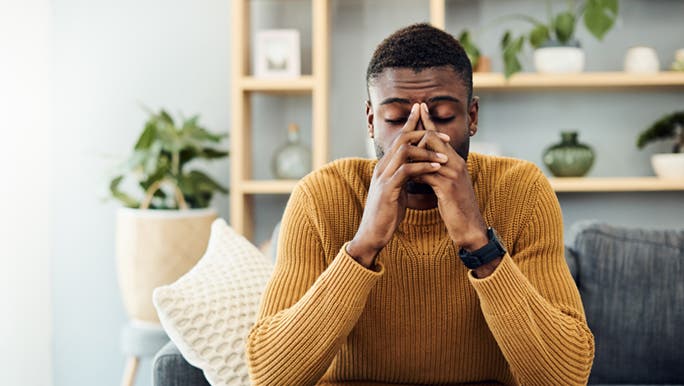 A man who’s wearing a yellow jumper, is sitting on the couch with both hands, resting on his face. He is showing signs of fatigue.