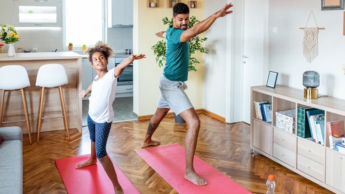 Two people are standing on exercise mats in their lounge room and stretching their arms to improve flexibility.
