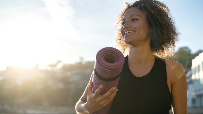 Smiling woman in black active wear holding a rolled up purple yoga mat