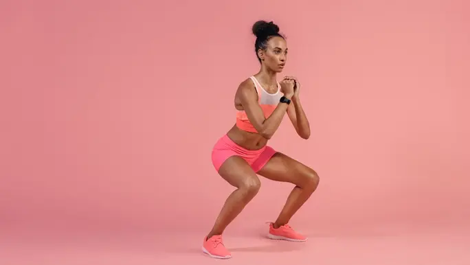 Young fit woman in pink active wear performing a squat exercise on a pink background