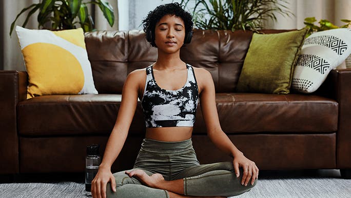 A woman meditates in front of a brown lounge with bright pillows. There are pot plants behind it. 