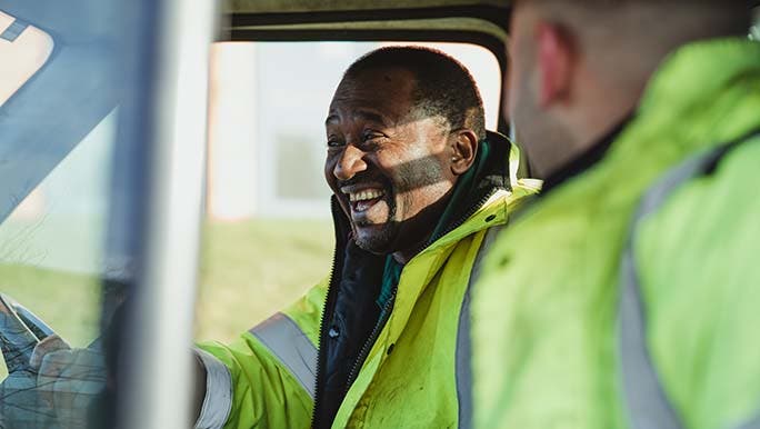 Two men are laughing and having a chat in a work vehicle because talking may help reduce work stress and the impact on relationships. 