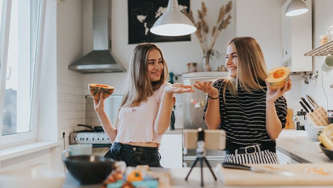 Two teenagers in a kitchen making a video, they each have half a pumpkin in their hands. 