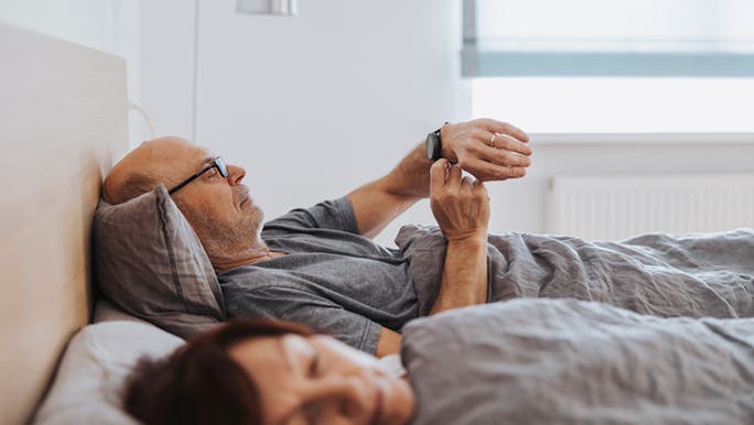 A senior man wearing glasses is lying in bed beside a sleeping woman and checking his smartwatch