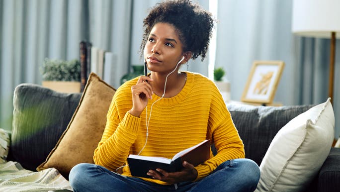A woman in jeans and a yellow sweater sits on her couch with a notepad and headphones thinking about 5 minute self care activities.