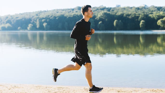 A man wearing all black is jogging next to a lake, he is enjoying the health benefits of running. 