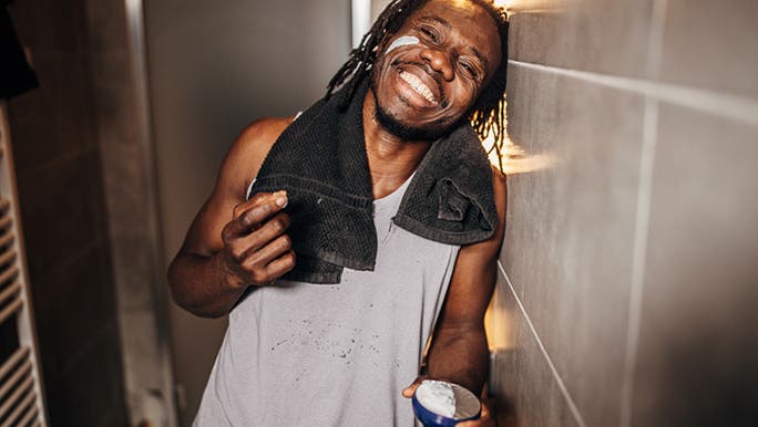 A young man is in the bathroom smiling as he applies face cream, which is part of a skin care routine