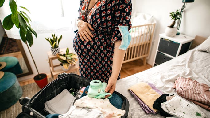 A pregnant woman is packing her hospital baby bag - it looks like she is ticking everything on the checklist. There is baby stuff all over the bed, folded neatly. 