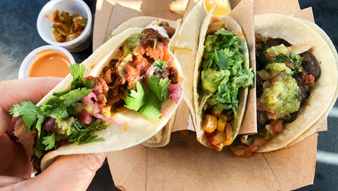 A hand holding some healthy looking takeaway tacos. They are full of veggies and herbs. 