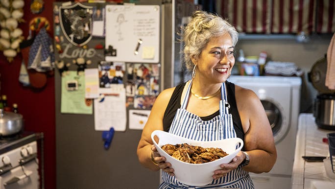 A smiling woman wearing a striped apron carries a bowl of food for a weekly family meal. 