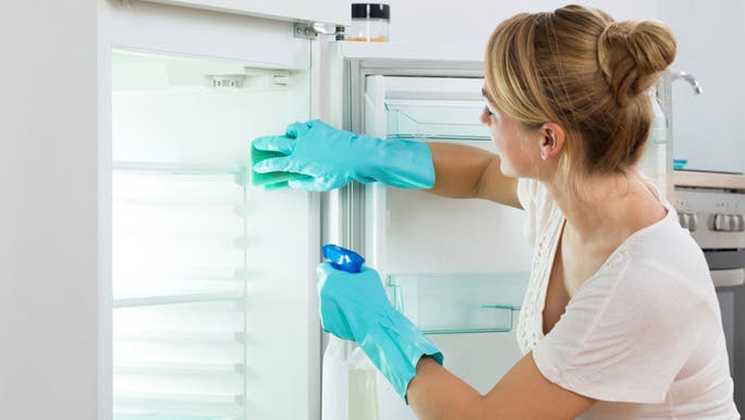 A young woman wearing a white T-shirt and wearing blue rubber gloves is cleaning out a fridge