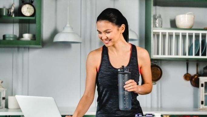 A fit young woman is smiling as she researches health supplements on a laptop sitting on a kitchen bench