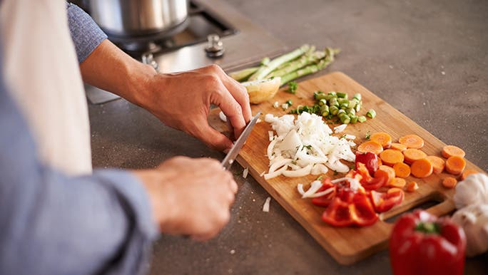 A man chops onions, carrots and capsicum on a wooden chopping board. He may be following a gut heath diet. 