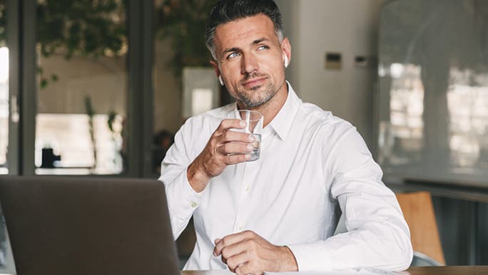 A businessman wearing a white shirt and sitting in front of a laptop is drinking a glass of water to get rid of a headache due to dehydration 