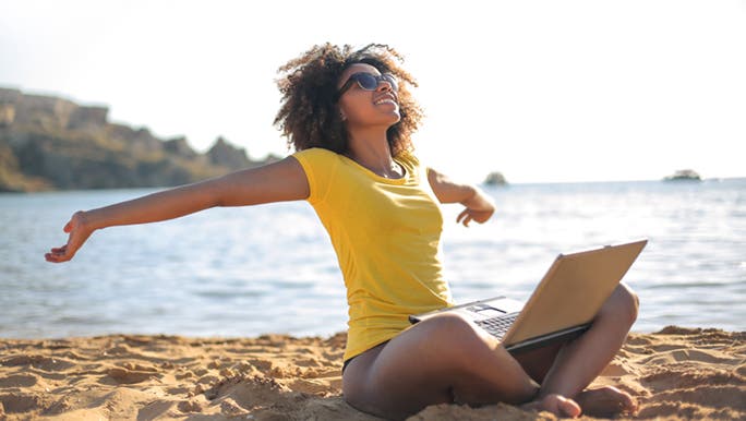 A woman in a yellow t-shirt sits at the beach, researching on her laptop how to get rid of frizzy hair.