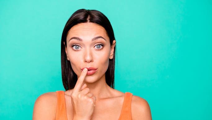 A young woman with an orange singlet stands in front of a blue wall with her finger to her lips, wondering how to get rid of dry lips.