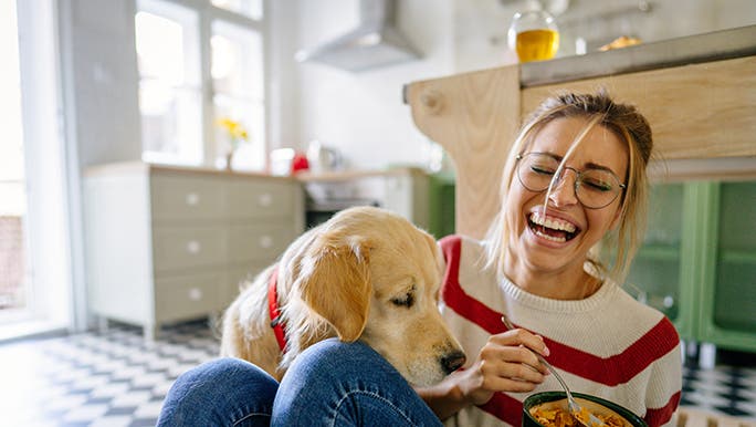 A lady sits on the floor of her kitchen eating something colourful out of a bowl. Her dog is trying to get in on the action. 