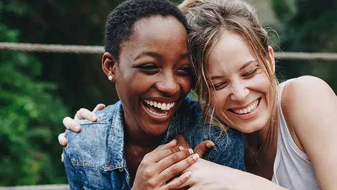 Two women smile while hugging each other in the outdoors.