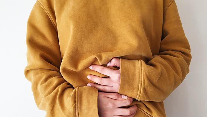 Up close, the torso of a woman wearing a mustard-coloured jumper is in the center of the frame. She is holding her gut in a way that suggests she’s experiencing bowel problems. 