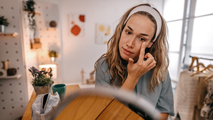 A young Caucasian woman is applying an eye cream in front of the mirror