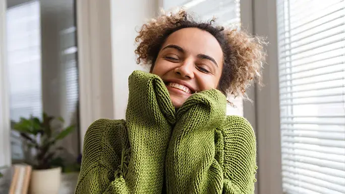 A woman in a soft green jumper smiles as she thinks about how to practise self-love. 