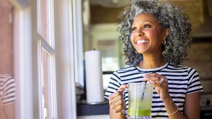 Woman with curly grey hair in a black and white striped tshirt sipping a green smoothie and looking out the window