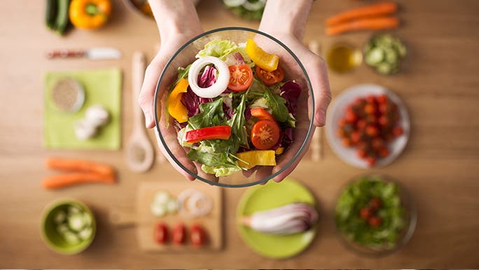 From above, a  glass bowl full of salad is being held above a table that is covered in vegan food. The background is blurred. 