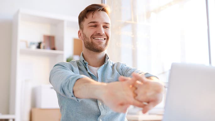 A man is stretching his arms and fingers while sitting at a computer desk. He is smiling and might be thinking about how good the stretch is feeling. 