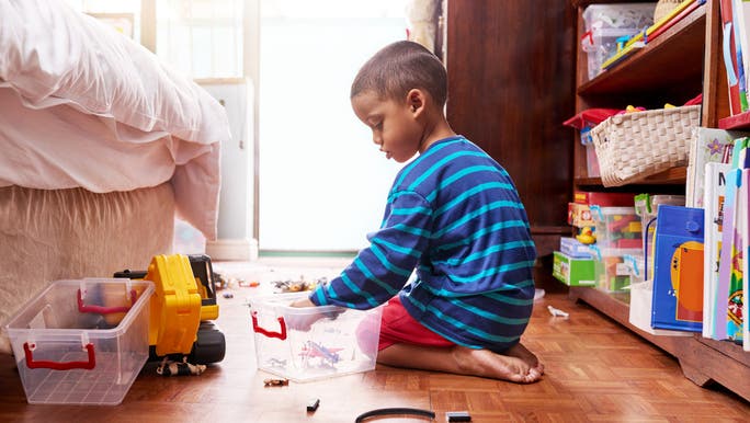 A young boy wearing a blue striped T-shirt is sitting on the floor of his bedroom playing with toys