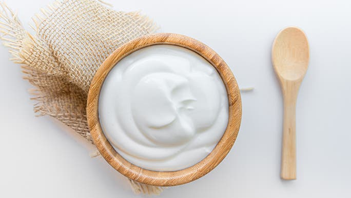 From above, a wooden bowl filled with yoghurt (a fermented food) is sitting on a white table next to a wooden spoon.  