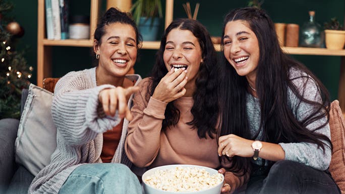 3 young ladies are sitting down eating popcorn, as they enjoy watching a movie. Some could say farts smell similar to popcorn.