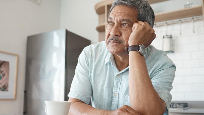 A senior man wearing a light blue shirt is leaning on a kitchen bench with his head on his hand with a sad expression on his face