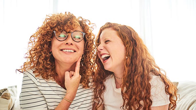 A mother and daughter with curly red hair sit together on a couch in front of a sunny window. 