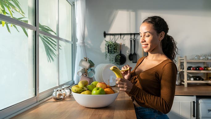 A woman with a dark complexion and wearing a brown top is peeling a banana, one of the best fruits for gut health