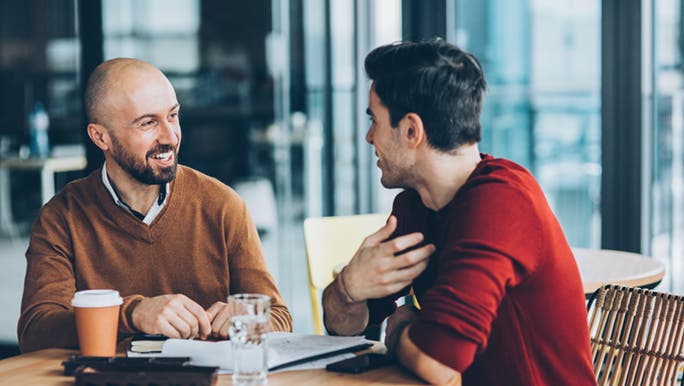 Two men sit at a cafe smiling while discussing how to say no to friends. 