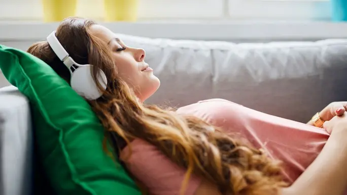 Young woman lying on the couch with a green cushion under her head. She is listening to headphones, relaxing with her eyes closed