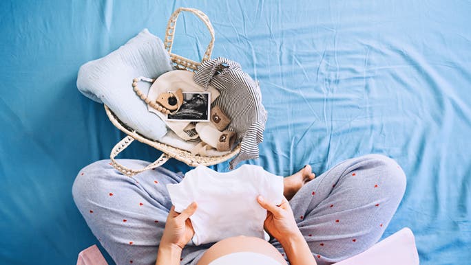 From above, a pregnant person sits cross legged on a bed with blue sheets. There is a baby basket with some rattles and ultrasound pics in front of her. She is folding a onesie carefully. 