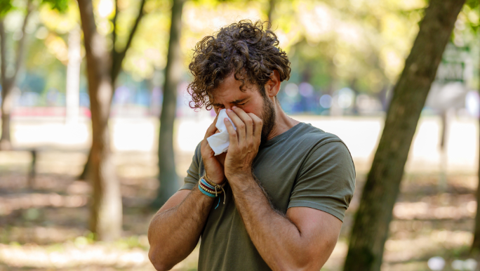A young man with allergies is standing in a park and blowing his nose