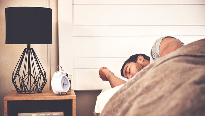 A dark-haired man is asleep in bed with a black lamp and white alarm clock on a bedside table beside him